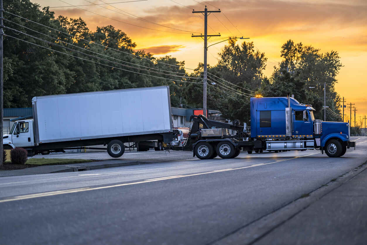 Blue heavy-duty tow truck hauling a broken semi with a box trailer on a local road.