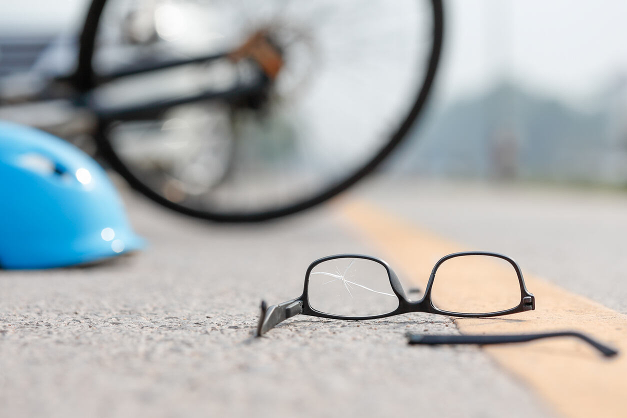 Broken eyeglasses and a bicycle helmet lying on the asphalt near a fallen bicycle, illustrating the severe impact and potential for brain and spine injuries in high-speed bicycle accidents.